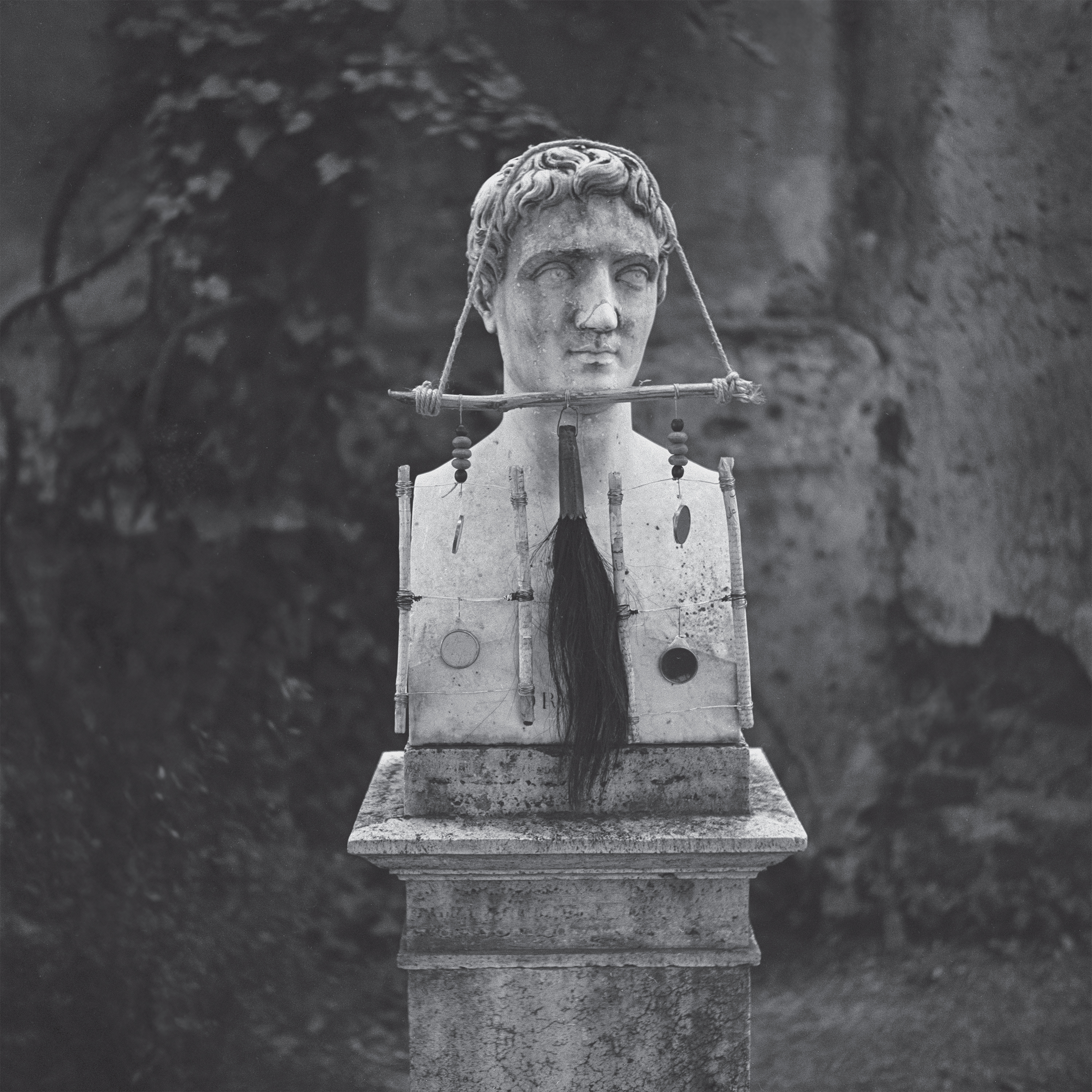 A weathered bust of a Roman figure, adorned with a long black hairpiece and mounted on a stone pedestal, set against a backdrop of aged walls and greenery.