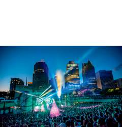 A large crowd surrounding a stage in the nighttime. Pink, blue, green and yellow lights emanate from the stage and the Detroit skyline is visible in the background.