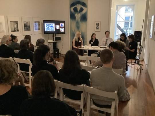 A panel of five people sitting at a table in the front of a room, with an audience facing them. There are artworks visible on the wall, and a screen in the corner of the room.
