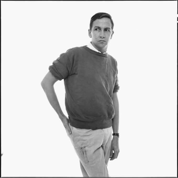 A black and white portrait of Robert Rauschenberg standing, looking into the distance, with his right hand on his hip. He is wearing a sweater with sleeves rolled up and light color pants. 