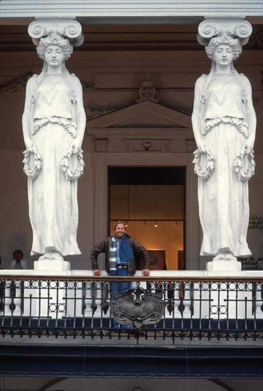 Robert Rauschenberg standing behind a balustrade, with two giant caryatids, columns carved in the shape of classical female figures, on either side of him.