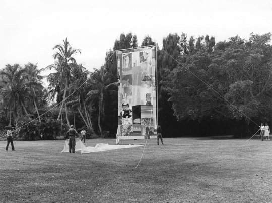 A large artwork stands upright in the middle of a grass field. A large piece of fabric, laying on the grass, is held on one side by a figure facing away from the camera. Multiple people stand at a distance holding ropes attached to the artwork's frame.