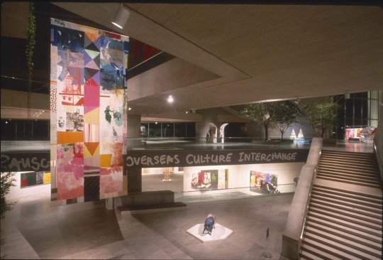 A two-story interior courtyard, with a large, colorful, banner-like artwork handing from the ceiling on the left, a large staircase on the right, and numerous, colorful artworks visible in the distance.