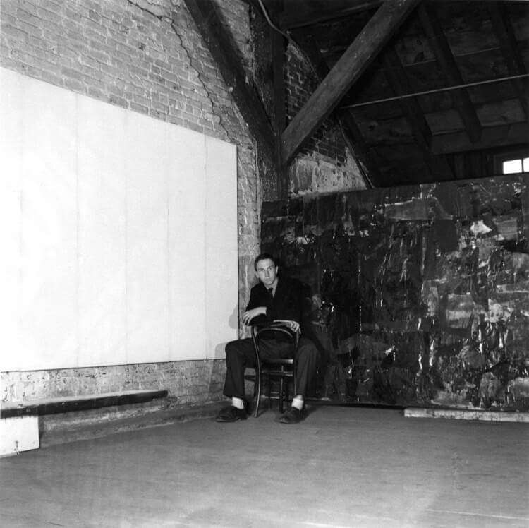 Artist Robert Rauschenberg sits backwards on a chair in the corner of his studio with a large white painting to the left and a large black painting to the right