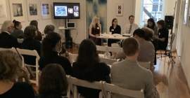 A panel of five people sitting at a table in the front of a room, with an audience facing them. There are artworks visible on the wall, and a screen in the corner of the room.