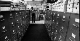 Robert Rauschenberg in a room full of file cabinets and boxes, leaning on an open file cabinet drawer and looking off to the side. 