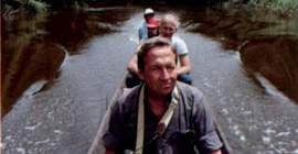 Robert Rauschenberg sits in a canoe, with a camera in his hands. Three people sit behind him. The canoe is in the middle of a river, surrounded by lush rainforest.