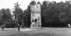 A large artwork stands upright in the middle of a grass field. A large piece of fabric, laying on the grass, is held on one side by a figure facing away from the camera. Multiple people stand at a distance holding ropes attached to the artwork's frame.