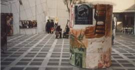 A large, colorful, cylindrical sculpture sits at the center of an outdoor courtyard. In the background, museum visitors sit at tables, while others look at artworks that hang on the courtyard walls.