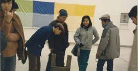 A group of people look down at a sculpture made of wood and tire tread that sits on the floor. A green, blue, and yellow fabric artwork is visible hanging on the wall in the background.