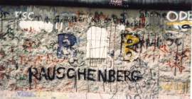 A view of a small section of the Berlin Wall. Among the largely illegible, multi-color graffiti is the name "Rauschenberg" in black, uppercase letters.
