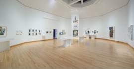 A panoramic view of a hexagonal room with framed artworks and vitrines along the white walls. Rauschenberg's large, vertical, three part print, Autobiography, is visible in the center.