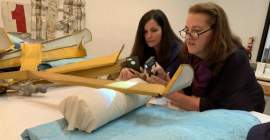 Two people examining part of a yellow, metal artwork. One is holding a shining a light, and the other is holding up a smartphone. The artwork is propped up on a white cushion and blue blanket.